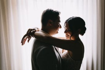 תמונה ובה אישה בשמלה לבנה מחבקת ומביטה בעיניים בגבר בחליפה אפורה. ברקע וילון לבן