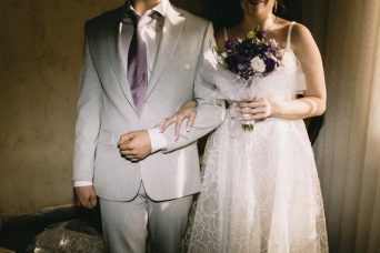 תמונה ובה גבר בחליפה אפורה ואישה בשמלה לבנה משלבים ידיים. האישה מחזיקה בזר פרחים בידה השניה